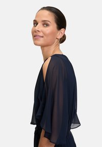 Mörkblå genomskinlig klänning med flödande ärmar, med ett urtag vid axeln och en bälte i midjan, visad på en modell från sidan.