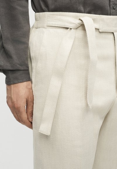 Κοντινό πλάνο σε μπεζ παντελόνι με ψηλή μέση και ζώνη από ύφασμα δεμένη σε κόμπο, φορεμένο με σκοτεινό μακρυμάνικο πουκάμισο, δείχνοντας ένα χαλαρό χέρι στο πλάι.