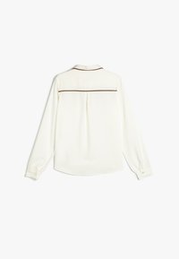 Camisa blanca de manga larga con un pliegue en la parte posterior, ribete marrón en el canesú y puños abotonados. Tejido suave con un corte relajado.