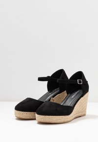 Schwarze Wildleder-Plateauschuhe mit einem mit Jute umwickelten Fußbett, ausgestattet mit einem Knöchelriemen und einer abgerundeten Zehenpartie. Glatte Textur mit minimalen Akzenten.