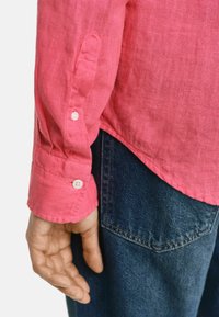 Camicia a maniche lunghe in lino rosa con polsini bottonati, abbinata a jeans in denim blu. Cuciture visibili e dettaglio della tasca sui jeans.