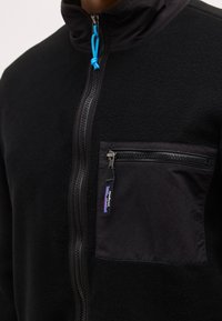 Μαύρο φλις μπουφάν με εμπρόσθιο φερμουάρ και τσέπη στο στήθος με φερμουάρ, που φέρει μια μικρή ετικέτα με το λογότυπο της Patagonia.