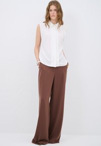 Witte mouwloze blouse met een knoopsluiting bij de kraag, gecombineerd met losse bruine broeken. Glad materiaal met een comfortabele pasvorm.