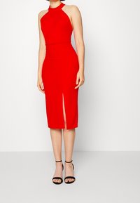 Femme portant une robe sans manches rouge à décolleté plongeant, avec une fente à l'avant, assortie à des sandales à talons hauts à brides de cheville noires.