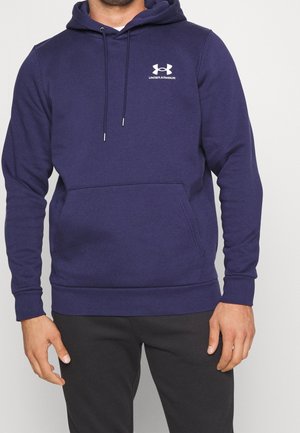 Mann trägt eine marineblaue Under Armour Kapuzenjacke mit Fronttasche und schwarze Hose, steht vor einem einfachen hellen Hintergrund.