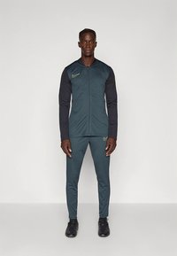 Športová súprava Nike v tmavozelenej farbe, so zapínacou bundou s čiernymi rukávmi a vyšívaným logom. Celý outfit dopĺňajú priliehavé nohavice.