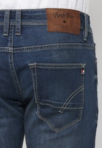 Tamno plavi traperice s kožnom oznakom na pojasu označenom "Kirsh Jeans." Imaju šavove na stražnjem džepu i malu oznaku brenda na džepu.