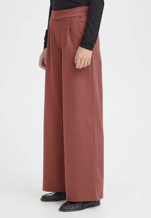 Woman wearing rust-colored wide-leg pants, black long-sleeve top, and black flat shoes, standing against a plain light background.