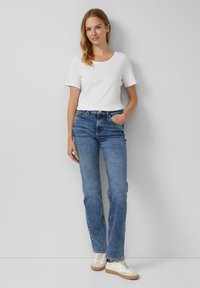 Weißes T-Shirt mit kurzen Ärmeln, kombiniert mit blauen Jeans im geraden Schnitt und weißen Sneakers mit braunen Akzenten, vor einem neutralen Hintergrund.