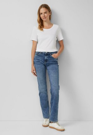 T-shirt blanc à manches courtes associé à un jean en denim bleu, coupe droite, et des baskets blanches avec des accents beiges sur un fond neutre.