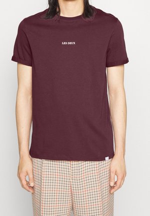 T-shirt en coton bordeaux à manches courtes, avec un logo blanc "LES DEUX" sur la poitrine. Associé à un pantalon à carreaux.