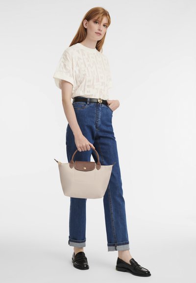 Beige handbag with brown leather accents, a structured shape, and a top handle, paired with a cream patterned t-shirt and blue jeans.