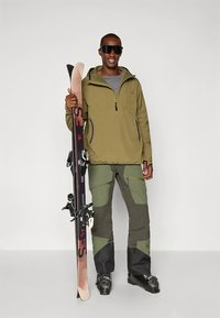 Équipement de ski comprenant une veste vert olive, une chemise grise et un pantalon vert-brun avec des accents renforcés. Skis à la main, lunettes de soleil noires portées.