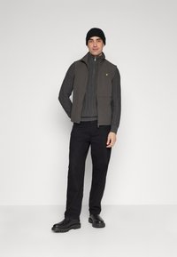 Gilet grigio senza maniche sopra un maglione grigio a costine, abbinato a pantaloni neri e stivali neri chunky. Un berretto completa il look.