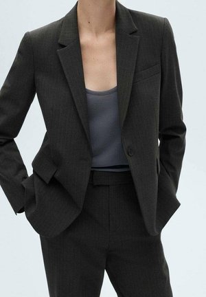 Person trägt einen dunkelgrauen Nadelstreifenanzug mit einem einreihigen Blazer und passenden Hosen, kombiniert mit einem grauen ärmellosen Oberteil.