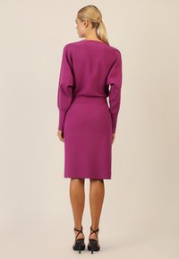 Vestido de punto acanalado fucsia con mangas largas y falda ajustada. Presenta un amplio escote barco y una cinturilla sin costuras, complementado con sandalias de tacón alto.