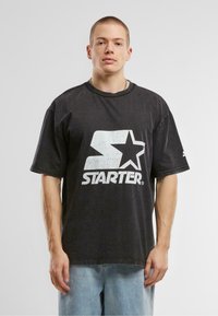 Juoda, per didelė polo marškinė su baltu grafiniu logotipu "STARTER", kuriame pavaizduota žvaigždė, trumpomis rankovėmis ir apvaliu kaklu, derinami su šviesiai mėlynomis džinsais.