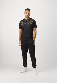 Mustat urheilupaidat, joissa on keltaisia yksityiskohtia ja aaltoilukuosi, yhdistettynä mustiin collegehousuihin, joissa on hienovarainen kuvio, ja valkoisiin lenkkareihin.