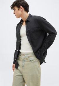 Νέος άνδρας που φοράει μαύρο πουκάμισο με κουμπιά πάνω από λευκή φανέλα και μπεζ τζιν, κοιτάζοντας προς τα αριστερά του, με φόντο ένα απλό σκηνικό.