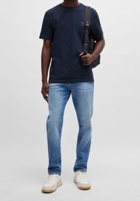 Man som bär en marinblå t-shirt, ljusblå jeans, vita sneakers och bär en svart ryggsäck på ena axeln mot en enfärgad bakgrund.