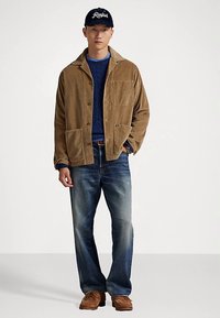 Ung mann iført en brun kordfløyelsjakke, blå kabelstrikket genser, falmede jeans, brune sko og en marineblå caps med "Ralph"-logo.