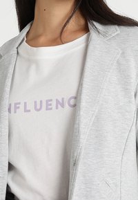 Světlě šedé sako s klopovým límcem přes bílé tričko s nápisem "INFLUENC" fialovou barvou. Hladká textury látky, střih na míru.