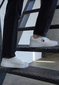 Zapatillas blancas con acabado de cuero liso, puntera redondeada y un logo decorativo en el lateral. Llevadas en una escalera negra, que muestra una textura escalonada.