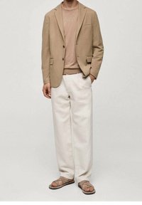 Blazer beige avec poches sur un pull léger beige, associé à un pantalon blanc ample et des sandales marron. Texture de tissu lisse partout.