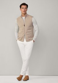 Gilet in maglia beige con colletto rialzato sopra una camicia grigia a maniche lunghe, abbinato a pantaloni bianchi e mocassini marrone chiaro, con chiusura a zip e tasche laterali.