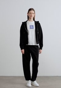 Μαύρο φούτερ με φερμουάρ, λευκό μπλουζάκι με μοβ τετράγωνο λογότυπο και μαύρη φόρμα. Λευκά αθλητικά παπούτσια, στέκεται σε ουδέτερο φόντο.