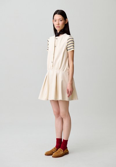 Vestido color crema con corsé ajustado y falda acampanada, complementado con una camiseta de manga corta a rayas. Incluye mocasines marrones y calcetines rojos.