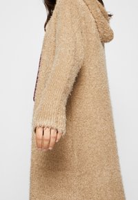 Beige neulottu neuletakki, jossa on pehmeä, pörröinen pinta, pitkät hihat ja huppu. Siinä on väljä istuvuus ja raakapäärmeiset hihansuut.