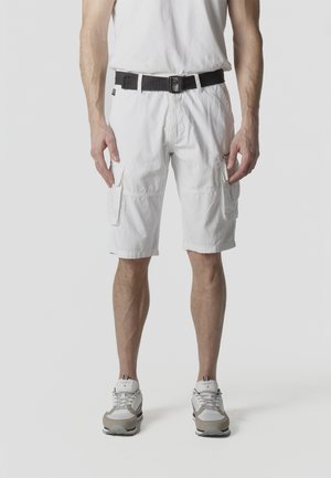 Hombre con pantalones cortos cargo blancos, cinturón negro, camiseta blanca y zapatillas grises y blancas, de pie frente a un fondo liso.