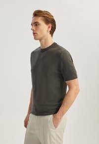 T-shirt a maniche corte in maglia verde scuro con collo rotondo e bordi a coste. Texture liscia, design aderente, abbinata a pantaloni beige chiaro.