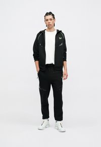 Melna zip-up hoodie ar neona zaļajiem akcentiem, apvienots ar melnajām sporta biksēm ar atstarojošiem elementiem. Modelis valkā baltas kedas.
