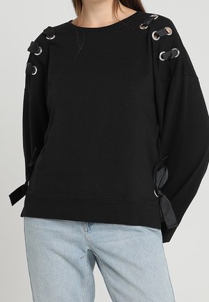 Zwart sweatshirt met lange mouwen, zilveren oogjes en zwarte linten gestrikt op de schouders en mouwen, gedragen met lichtblauwe jeans.