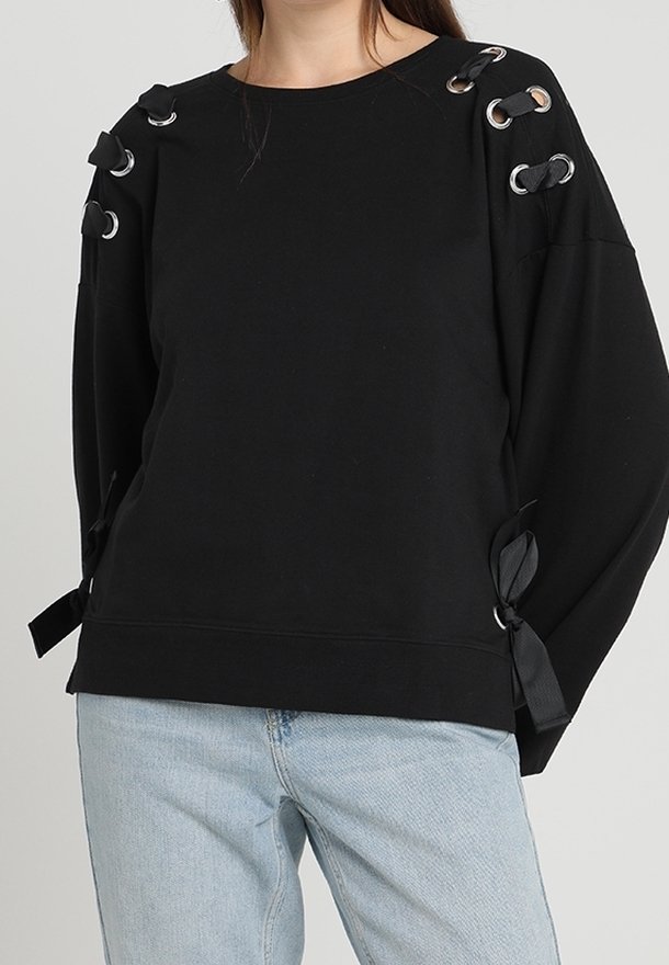 Sweat-shirt noir à manches longues avec œillets argentés et rubans noirs noués sur les épaules et les manches, porté avec un jean bleu clair.