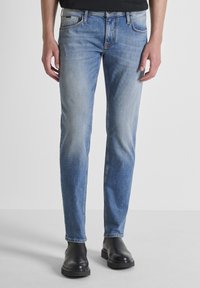 Svijetloplave traperice slim kroja, standardnog dizajna s pet džepova, s blagim strugotinama sprijeda. Uparene s crnim čizmama.