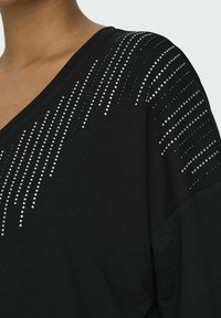 Top nero a V con maniche corte, caratterizzato da una gradazione di strass argento sulle spalle, creando un motivo diagonale. Tessuto morbido.