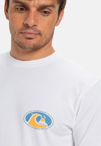 Hvit bomulls T-skjorte med et fargerikt grafisk logo i blått, gult og oransje, plassert på venstre brystområde. Korte ermer, rund hals.