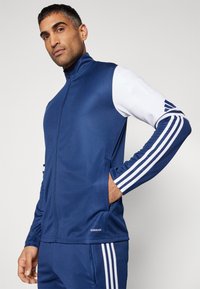Marineblauwe zip-up jas met witte accenten, voorzien van gestreepte mouwen en verborgen zakken. Gemaakt van een gladde, ademende stof.