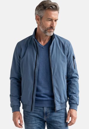 Mann mit grauem Haar und Bart, der eine blaue Bomberjacke mit Reißverschluss, einen blauen V-Ausschnittpullover und blaue Jeans trägt und nach unten schaut.