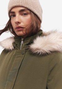 Parka verde oliva con cappuccio foderato in pelliccia sintetica, dotata di chiusura a zip, bottoni a pressione e un berretto beige lavorato a maglia.
