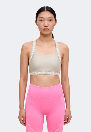 Γυναίκα που φοράει μπεζ αθλητικό σουτιέν Under Armour και ροζ κολάν με ψηλή μέση, στέκεται μπροστά σε ένα απλό φωτεινό φόντο.