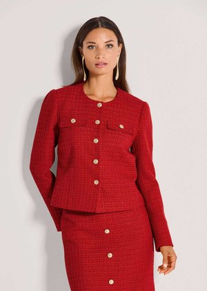 Vrouw draagt een rood gestructureerd jasje met knopen en bijpassende rok, met lange oorbellen, staand tegen een effen lichte achtergrond.