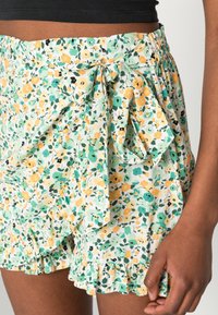 Blommiga omlottshorts i grönt, gult och svart med veckad fåll och rosett i midjan, tillverkade av lättviktsmaterial.