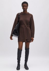 Bruine gebreide jurk met lange mouwen, strik in de taille, textiel met structuur, gecombineerd met zwarte kniehoge laarzen en een zwarte tas.