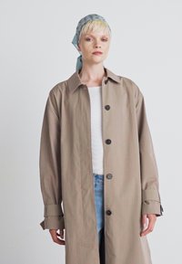 Jasnobrązowy płaszcz trench z prostym krojem, z przodu z guzikami i szerokimi rękawami. Noszony na białej bluzce i niebieskich dżinsach, uzupełniony jasnoniebieskim szalikiem.