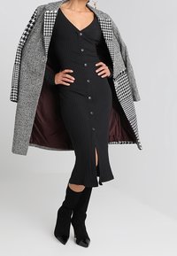 Svart ribbad midi-klänning med knappdetaljer och en framskjutning, överst i kombination med en grå hundtandsmönstrad kappa. Svarta stilettklackar fulländar looken.