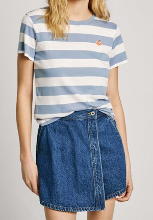 Jeune femme portant une chemise à manches courtes à rayures bleu clair et blanc et une jupe en denim bleu taille haute, debout avec les bras détendus.